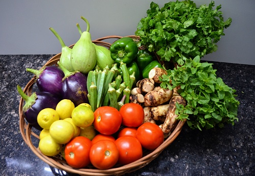 Cane basket full of vegetables. Red tomatoes, round bottle gourd brown-cream taro, purple eggplant, green bell peppers, yellow lemons, green ladyfingers, green coriander bunch and green mint leaves.