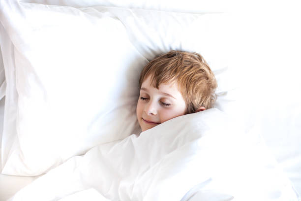 Sleeping little boy lying in bed under white blanket. Smiling boy with closed eyes relaxing in bedroom. Healthy sleep, sweet dreams concept. Copy space. Selective focus.