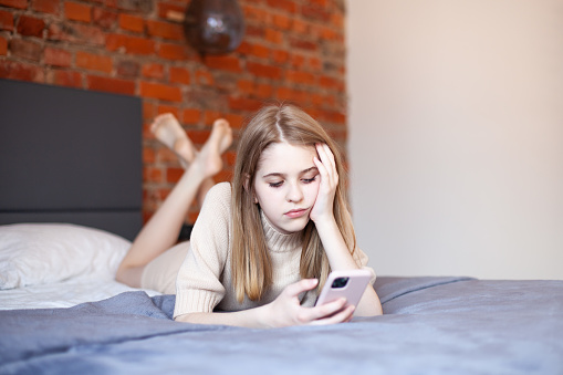 Thoughtful teen girl lying on bed with smartphone in hand. Teens and gadgets, online courses for teens, online shopping, mental health and relationship problems of teenagers. Selective focus.