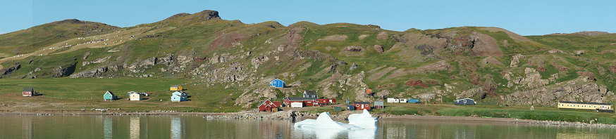 Panoramic view of living in West Greenland with the sea, the mountains, the green fields, the multi-coloured wooden houses, the icebergs floating around in the sea; summer atmosphere and sun