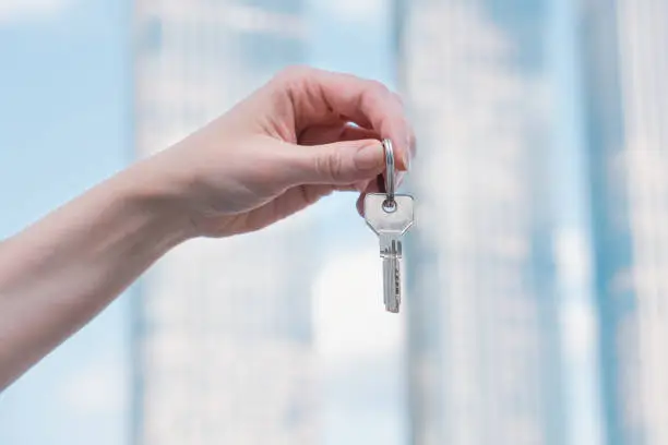 Woman's hand holds a key against the backdrop of a modern multi-storey building with a glass facade. The concept of buying residential real estate, winning the lottery, affordable mortgage