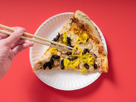 Human hand picking at pizza slice on paper plate using chopsticks. Humorous and unusual juxtaposition of traditional pizza being eaten using an Asian utensil. On red background with copy space.