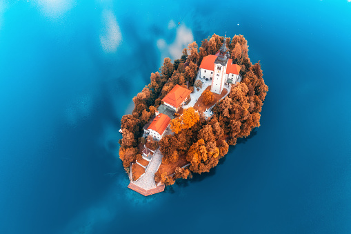 Aerial view of beautiful church and buildings on the island on the famous Bled Lake, Slovenia in autumn morning. Top drone view of chapel, houses, orange trees, blue water in fall. Landscape. Travel