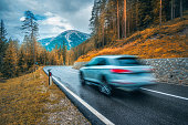 Blurred car in motion on the road in orange forest in rain in autumn. Dolomites, Italy. Perfect mountain road in overcast day in fall. Roadway, pine trees in alps. Transportation. Highway in woods