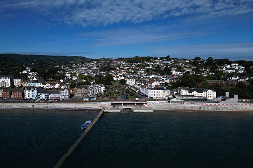 The drone gives a high angle view of the town of Dawlish showing: a boat moored at Dawlish Quayside; Dawlish Railway Station with its in-progress, new bridge; the modern concrete sea wall defence barrier; traffic through the town centre and seafront properties, hotels and guesthouses. Dawlish is a popular UK holiday resort and destination known for its native population of Black Swans on Dawlish Water. The railway line at Dawlish was notorious for frequent flooding during high tides and storms, which would often delay or halt train services from Exeter to Cornwall.