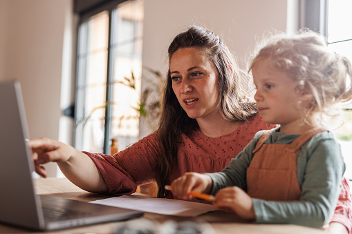 Portrait of mid adult woman sitting at table, trying to work from home, caring for her adorable toddler son who is sitting in her lap and distracting her.