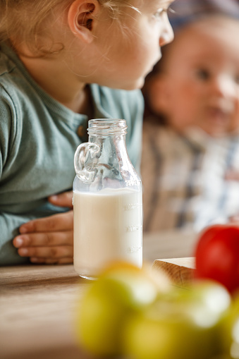 Close up shot of a glass bottle filled with milk on a table with baby boy and his toddler brother in the background.