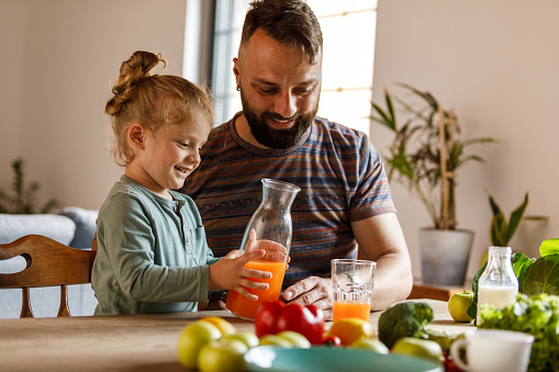 Portrait of cute little toddler and his father pouring fresh orange juice in a glass together at the dining table.