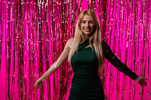 Front view of cool young blond woman walking through a shiny pink tinsel curtain. She is looking at camera with confidence.