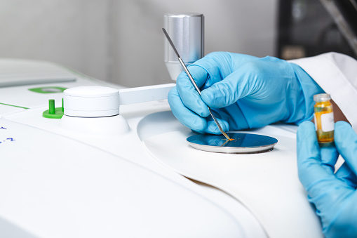 Hand of scientist using a stainless spatula to scoop sample solid from vial to the Fourier Transform Infrared Spectroscopy FTIR pedestal instrument for substance analysis and chemical characterization.