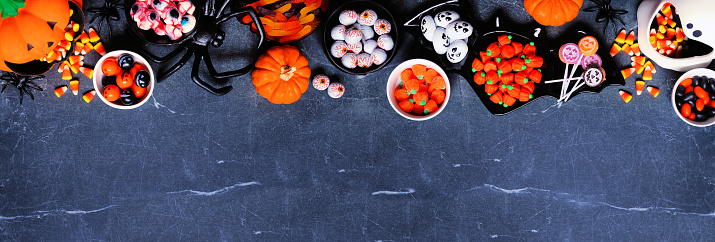 Halloween trick or treat candy top border. A selection of spooky sweets. Overhead view on a dark stone banner background with copy space.