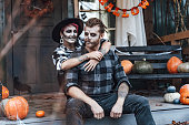 Scary love family couple man, woman celebrating halloween.Terrifying black skull half-face makeup,witch costumes,tattoo, stylish images,jacket,hat.Horror,fun at photoshoot, holiday party. Decorating of porch. Home, outdoor,street pumpkin jack-o-lantern