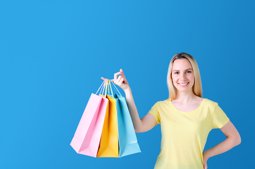blonde girl smiling standing in yellow t-shirt with paper shopping bags in one hand and the other resting on hip over blue background