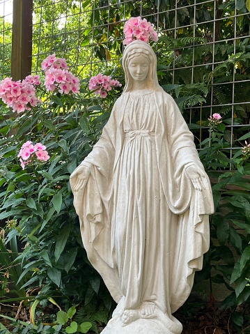 Mother Mary Statuary in garden with flowers. Pink phlox blooms and green leaves surround her as she stands in front of the garden fence.
