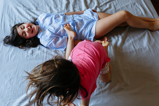 one smiling happy girl in blue pajamas lying down on the bed and other child sister jumping over