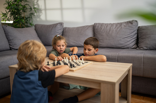 Cheerful kids, two brothers and sister sitting on floor and playing chess in living room at home