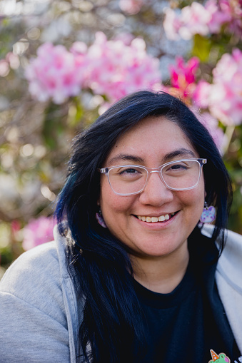 Portrait of a young woman with dark blue dyed colored hair smiling at the camera. She is standing next to some flowers in full bloom. Shot in the spring time in Portland Oregon.