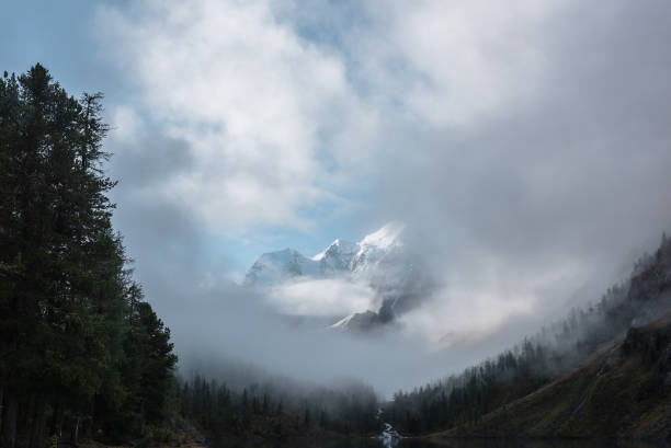 istock Tranquil scenery with snow castle in clouds and alpine lake. Mountain creek flows from forest hills into glacial lake. Snowy mountains in fog clearance. Small river and coniferous trees in low clouds. 1641115453