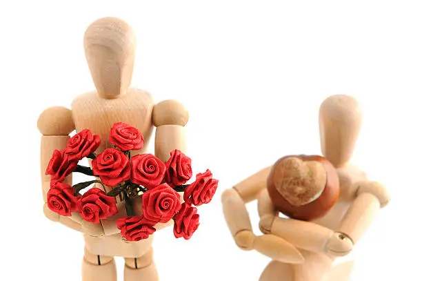 Wooden Mannequins showing their love. Situation of boy asked girl to engaged to be married. right woody kneeing in front of her. she holding roses in her arms and he a chestnut with heart shape to give her his heart. focus on froses. perfect valentines day background.