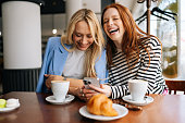 Portrait of two cheerful beautiful women friends sitting in cafe indoors, looking at phone and talking, laughing. Happy pretty girlfriends using social media