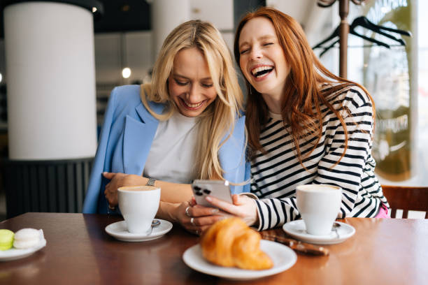 portret dwóch wesołych pięknych przyjaciółek siedzących w kawiarni w domu, patrzących na telefon i rozmawiających, śmiejących się. szczęśliwe ładne dziewczyny korzystające z mediów społecznościowych - coffee buzz zdjęcia i obrazy z banku zdjęć