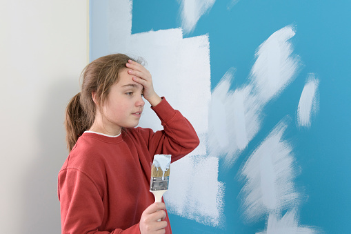 Teenage girl has headache at first time paints the wall. Girl painting a wall with a paint brush. Lifestyle