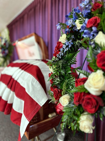 Selective focus veterans wreath of blue Delphinium with red and white roses near open casket draped with American flag in viewing parlor.