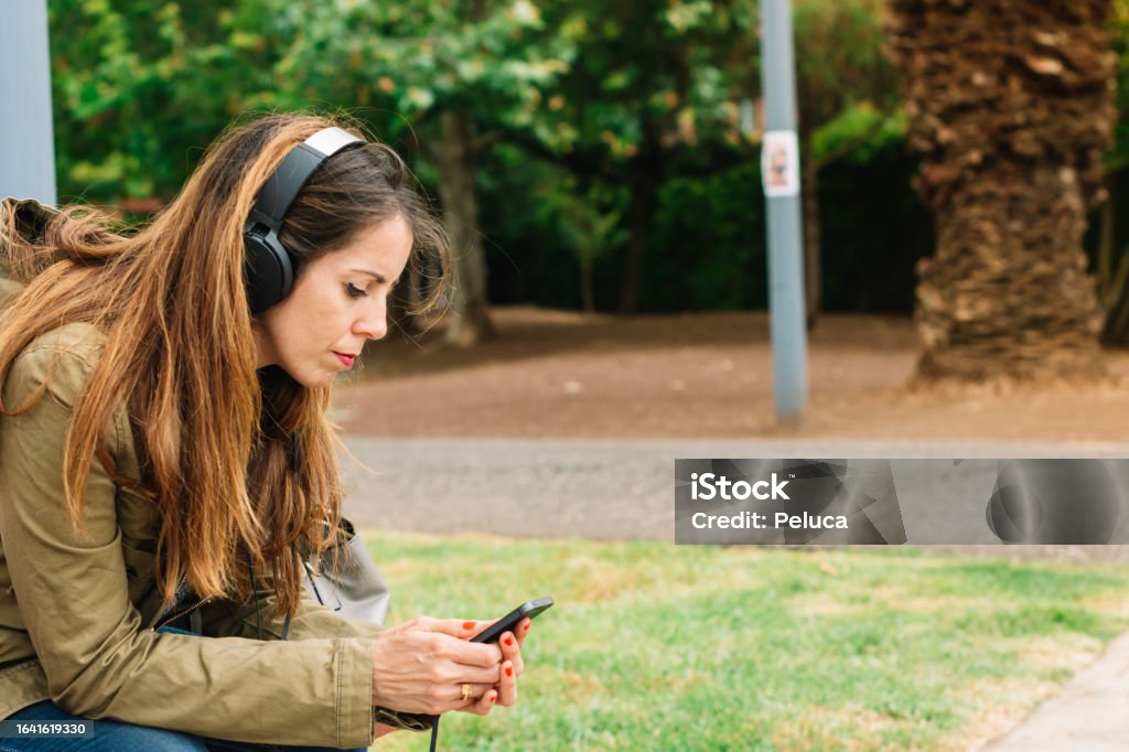 Woman listening to music with headphones and watching her cell phone in a park Music Streaming Service Stock Photo