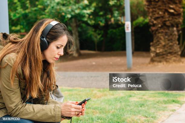 Woman Listening To Music With Headphones And Watching Her Cell Phone In A Park Stock Photo - Download Image Now