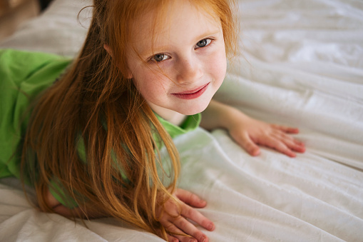 A red-haired little girl in a green T-shirt lies on a white bed at home and smiles. Portrait of a five year old girl.