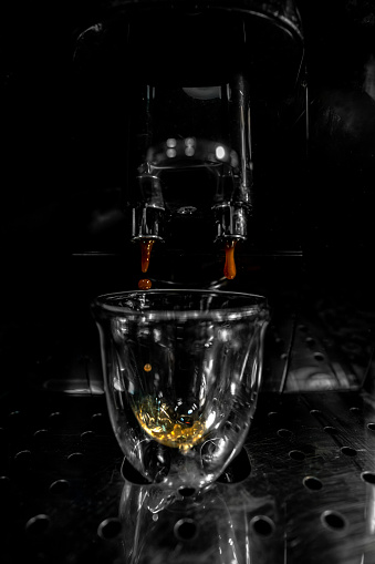 First drops of coffee falling through the double spout into a glass cup with foam and bubbles from an automatic espresso machine with a black and white background