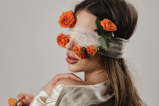 Portrait of a beautiful young woman having covered eyes with roses and cotton bandage, studio shot in front of white background