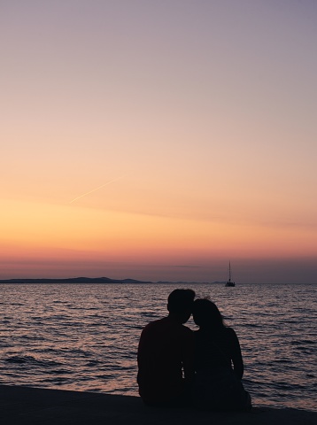 A vertical of a romantic couple sitting against the sea at sunset