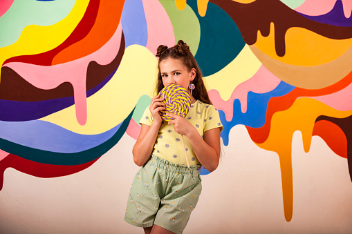Portrait of cover child girl covering mouth with giant round lollipop at colorful wall, looking at camera. Lovely kid in yellow t-shirt poses with candy on stick. Summer sweet concept. Copy text space