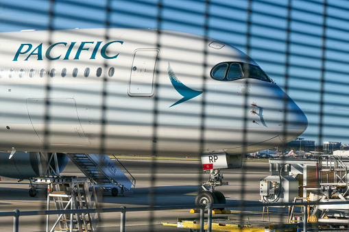 The forward section of a Cathay Pacific Airbus A350-941, registration B-LQG, parked at Gate 73 of Sydney Kingsford-Smith Airport near the fire station. In the background is the international terminal. This image was taken through a steel security fence from Kyeemagh Avenue, Kyeemagh on windy, cold and sunny afternoon close to sunset on 20 August 2023.
