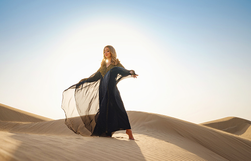 Desert adventure. Young arabian Woman posing in traditional Emirati dress abaya in sanddunes of UAE desert at sunset. The Dubai Desert Conservation Reserve, United Arab Emirates.