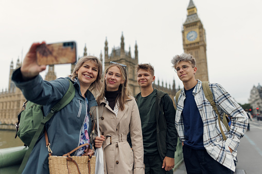 Family sightseeing London, UK. Overcast summer day. Mother and three teenagers are taking selfies on the Westminster bridge in front of the Big Ben.\nCanon R5