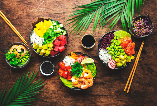 Poke bowl set with tuna, salmon, shrimp, avocado, mango, white rice and other ingredients. Soy sauce and sesame seeds. Rustic wooden table background, top view