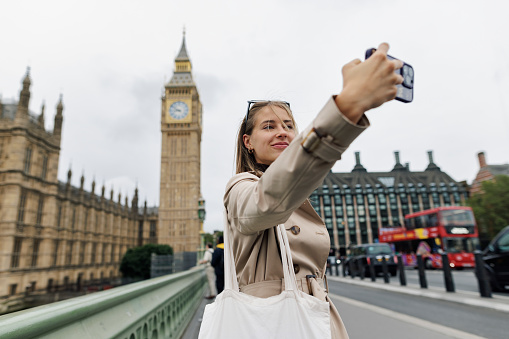 Teenage girl sightseeing London, UK. Overcast summer day. The girl is standing in front of the Big Ben and taking photos with a smartphone.\nCanon R5
