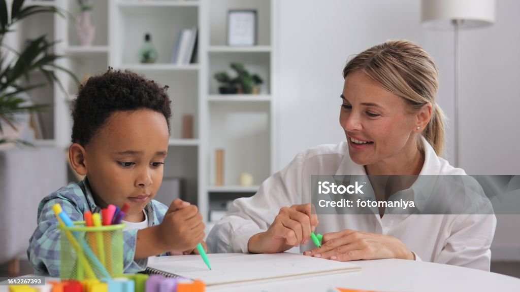 Adorable little african american boy with curly hair having fun at pediatric specialist appointment, happy child drawing with female therapist, exercises for children with autism. Child Stock Photo