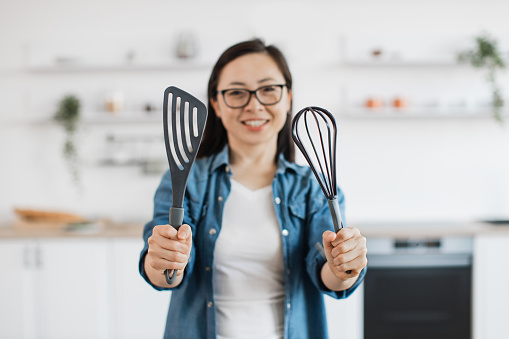 Focus on black whisk and spatula held by happy japanese lady in denim outfit standing on room background. Smiling housewife deciding on kitchen utensils before practising cooking skills at home.