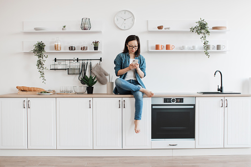 Full length view of asian woman using smartphone while sitting on countertop in white spacious kitchen. Carefree barefoot lady searching for new recipes on culinary websites using digital device.