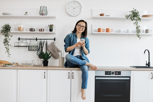 Happy smiling woman in casual wear typing on mobile phone while relaxing in kitchen interior. Joyful asian adult downloading audiobook for entertainment from online library using digital device.