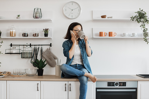 Young asian lady with cell phone holding grey mug while taking seat on countertop of stylish kitchen. Focused healthy female in glasses talking with friend on gadget while drinking morning coffee.