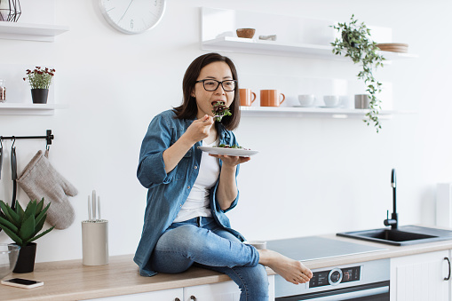 Young asian cook tasting vegetarian dish while sitting with one ankle over another knee in kitchen interior. Healthy slim lady in glasses creating homemade meal from scratch on sunny weekend.