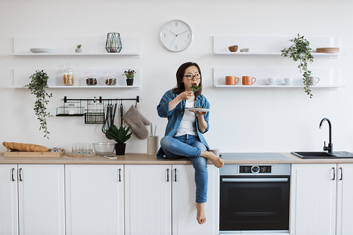 Full length view of attractive woman resting on countertop with vegetarian lunch on plate in stylish kitchen. Good-looking asian lady nibbling at salad loaded with health benefits of leisure time.