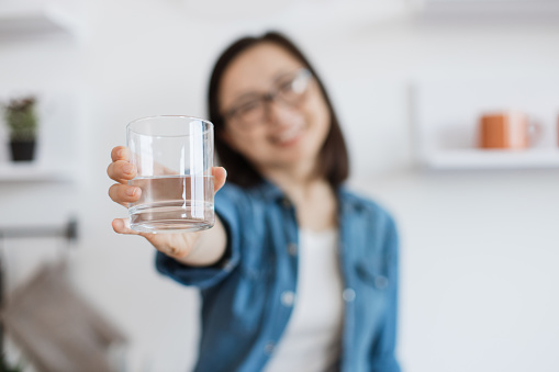Focus on tumbler glass half-full with clean liquid being reached out by happy asian lady in casual wear. Cheerful housewife encouraging to getting well-hydrated for improving health and mood.
