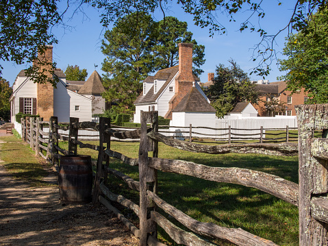 A wooden fence and barrel leading to white colonial houses beneath a clear blue sky in Williamsburg, Virginia.