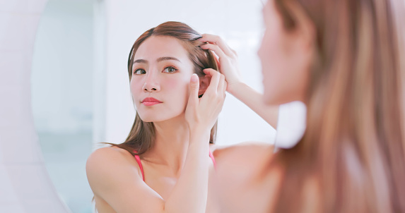 close up asian woman standing in front of mirror is checking her hairline inside bathroom at home