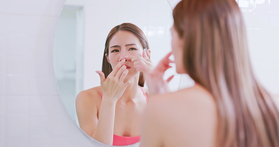 close up side view asian woman standing in front of mirror has bad unhealthy skin and touching her face inside bathroom at home - she squeeze pimple